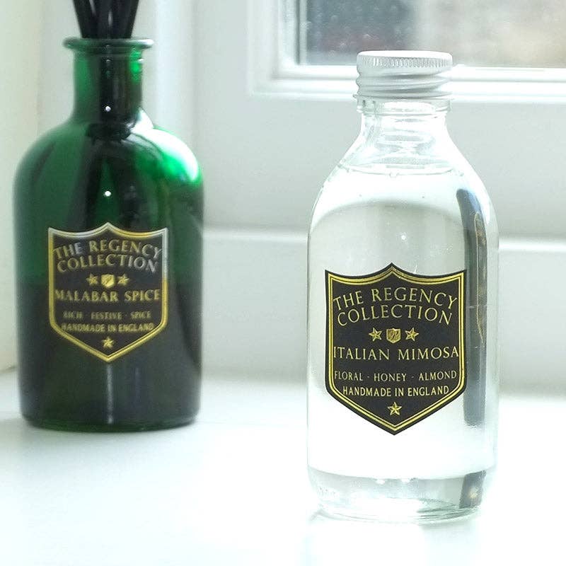 Two decorative bottles on a white surface near a window: a dark green glass reed diffuser bottle labeled 'The Regency Collection Malabar Spice' and a clear glass bottle labeled 'The Regency Collection Italian Mimosa' with white liquid and a silver cap.